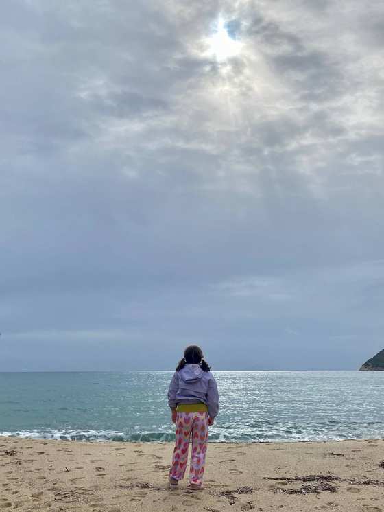 Una bambina di spalle sulla sabbia guarda il mare sotto un cielo coperto con un raggio di sole